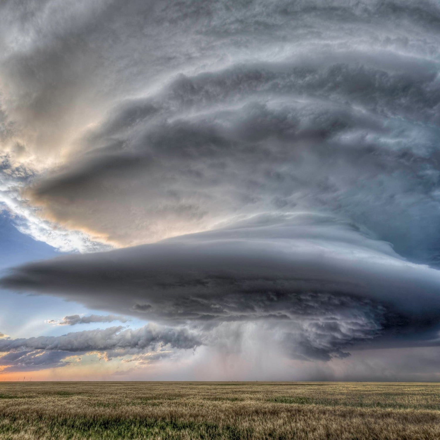 A massive, multi-layered storm cloud formation above an open field, with a gray sky and hints of sunlight on the horizon.