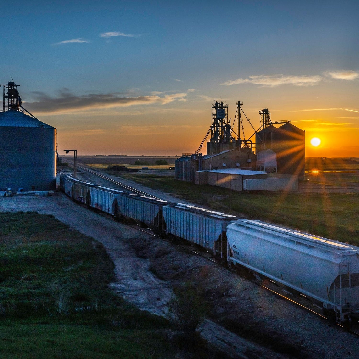 Train passing by grain silos at sunset in a rural area, with a clear sky and visible sun near the horizon.