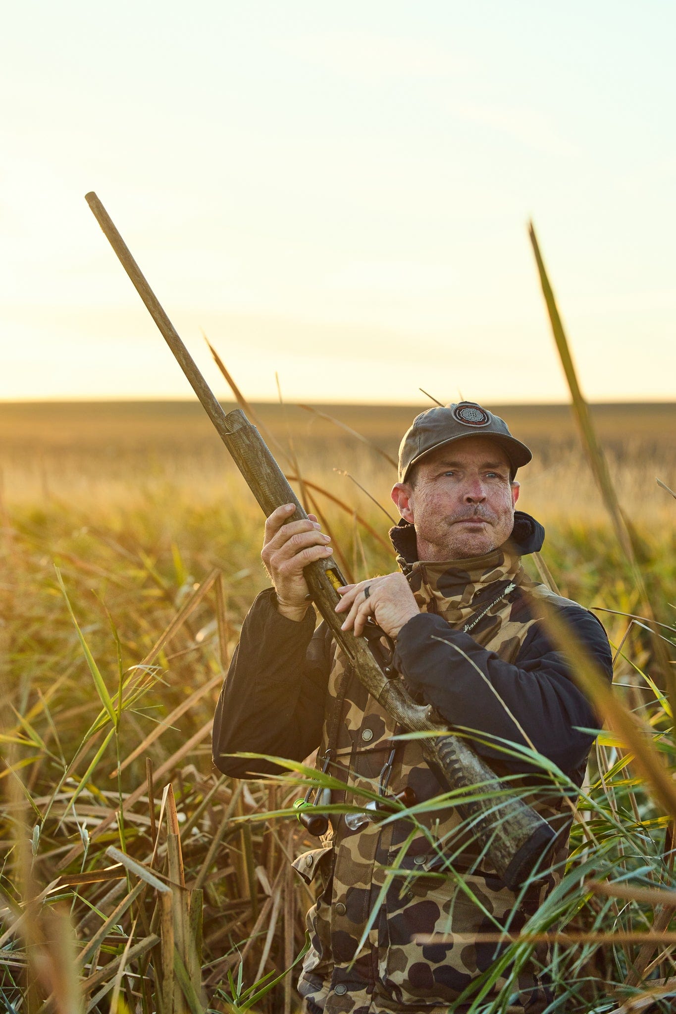 Person in camouflage, holding a rifle, stands in a grassy field during daylight.