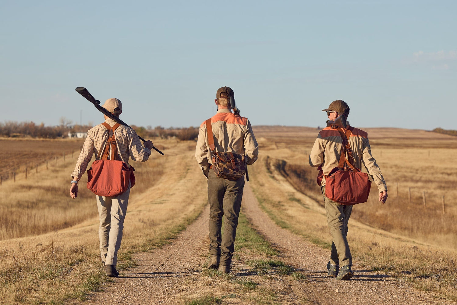 Three people in outdoor gear and carrying bags walk down a rural dirt path flanked by open fields under a clear sky.