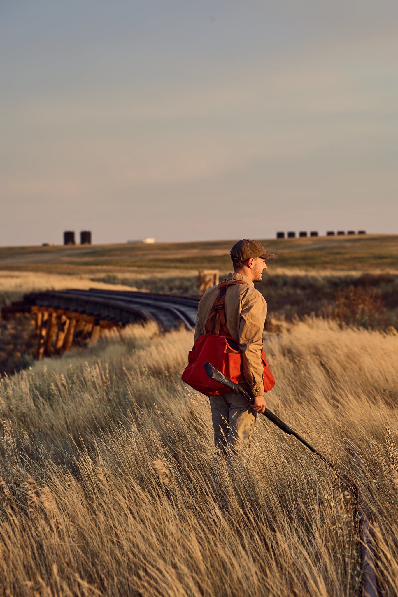 A person stands in a grassy field, holding a rifle and wearing a red bag, looking towards a distant bridge under a cloudy sky.