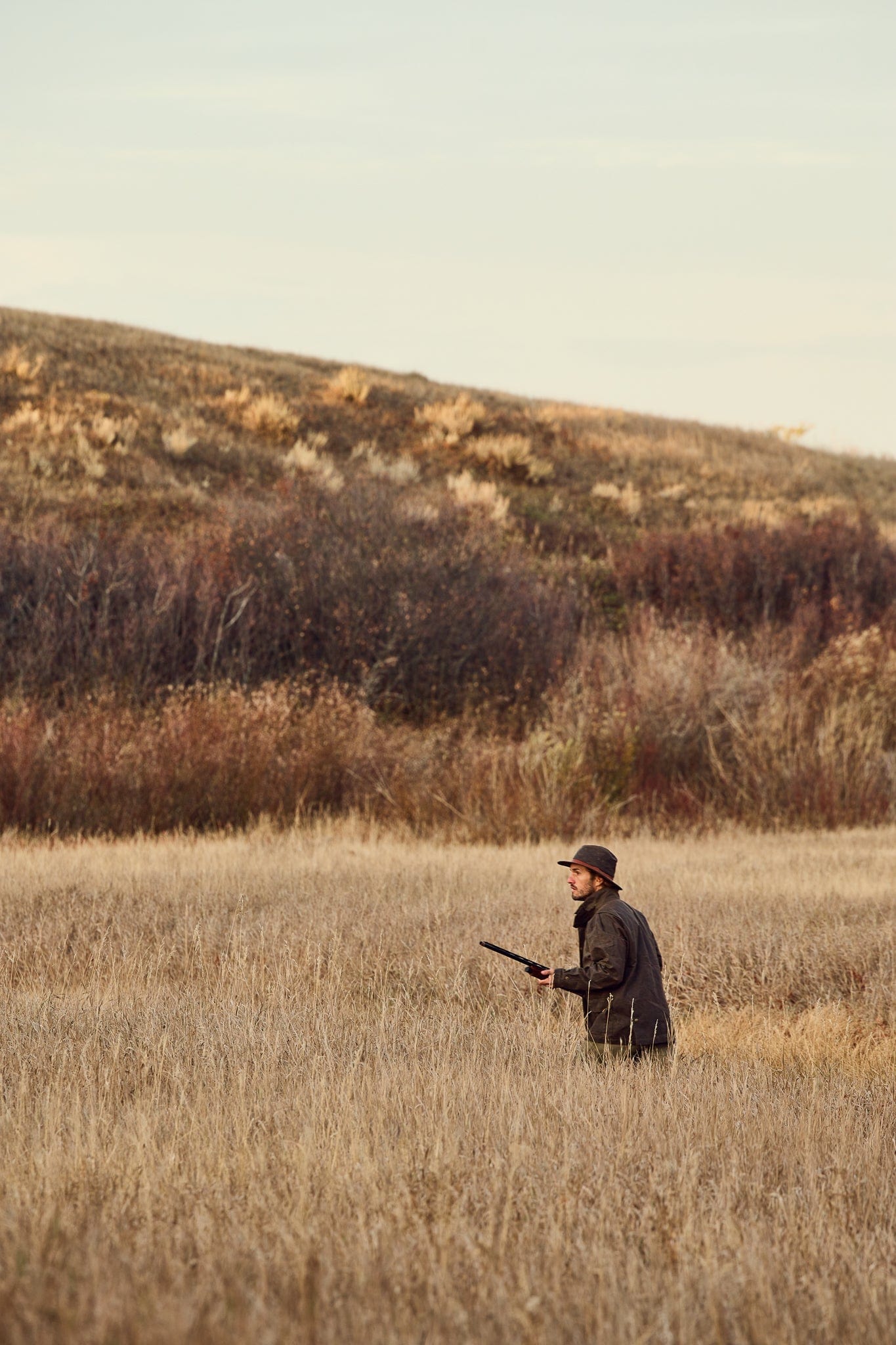 Person wearing a hat and holding a rifle, walking through a tall grassy field with hills and shrubs in the background.