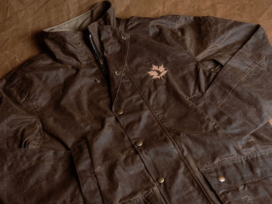 A brown waxed jacket with snap buttons and a maple leaf emblem on the chest, laid flat on a brown surface.