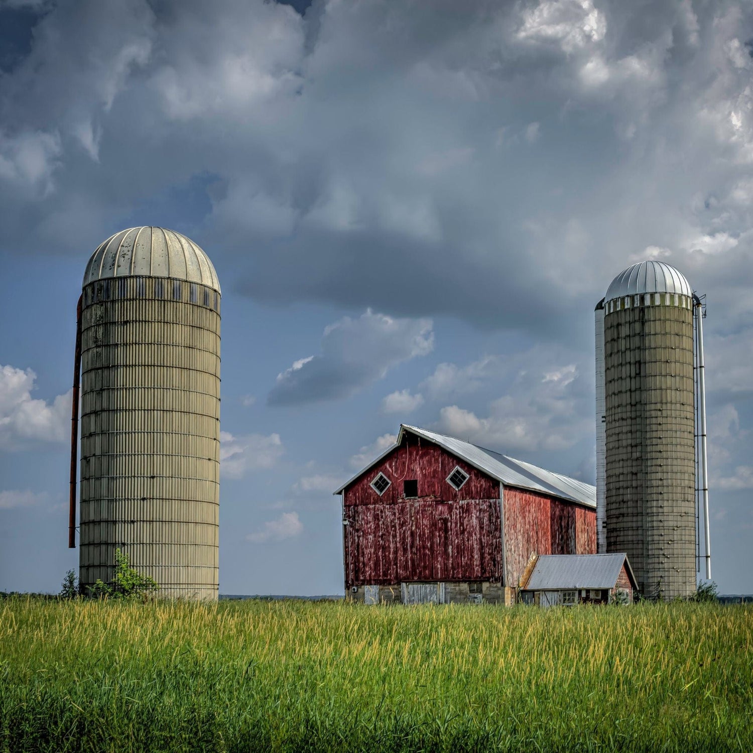 Red barn with two silos in a grassy field under a cloudy sky.