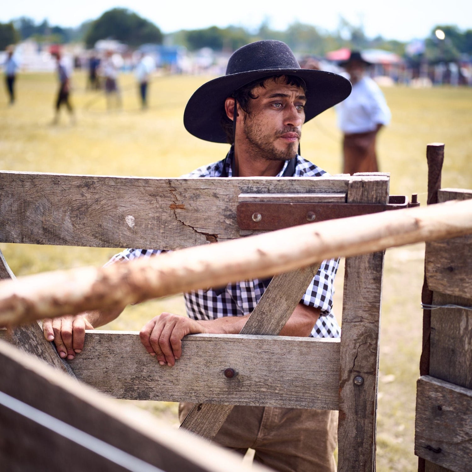 Man in a checkered shirt and black wide-brimmed hat leans on a wooden fence at an outdoor event with people in the background.