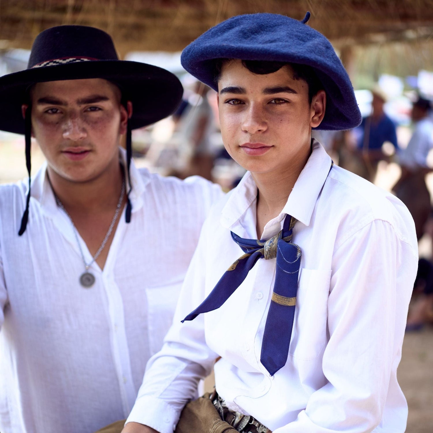 Two people in traditional attire with hats and scarves, standing in an outdoor setting with blurred figures in the background.