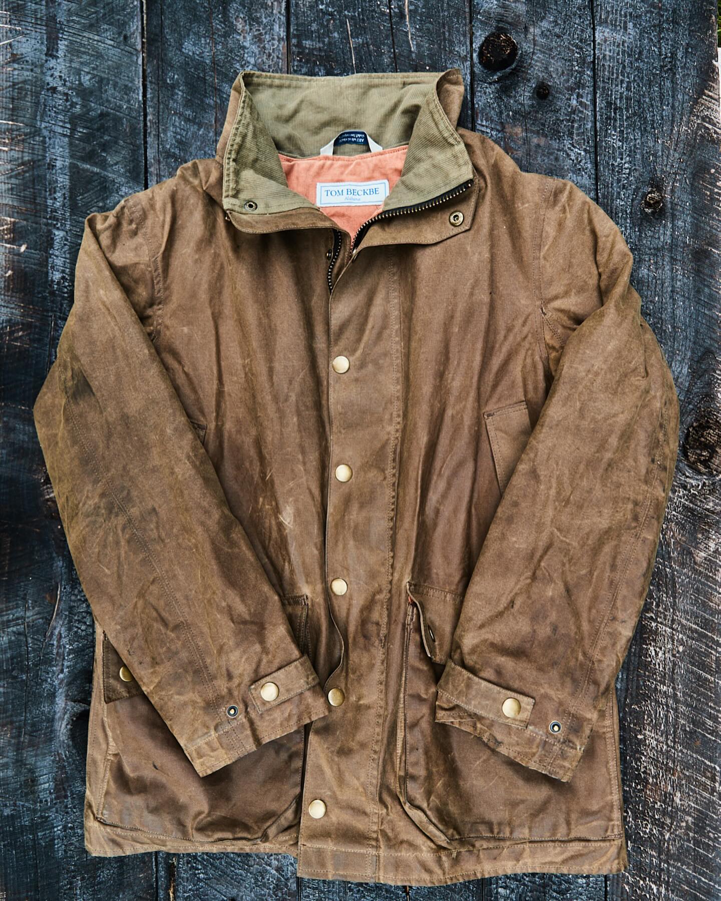 Brown waxed canvas jacket with button closures and a high collar, laid on a dark wooden surface.