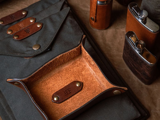 A leather valet tray and flasks are displayed on a brown surface with canvas pouches in the background.