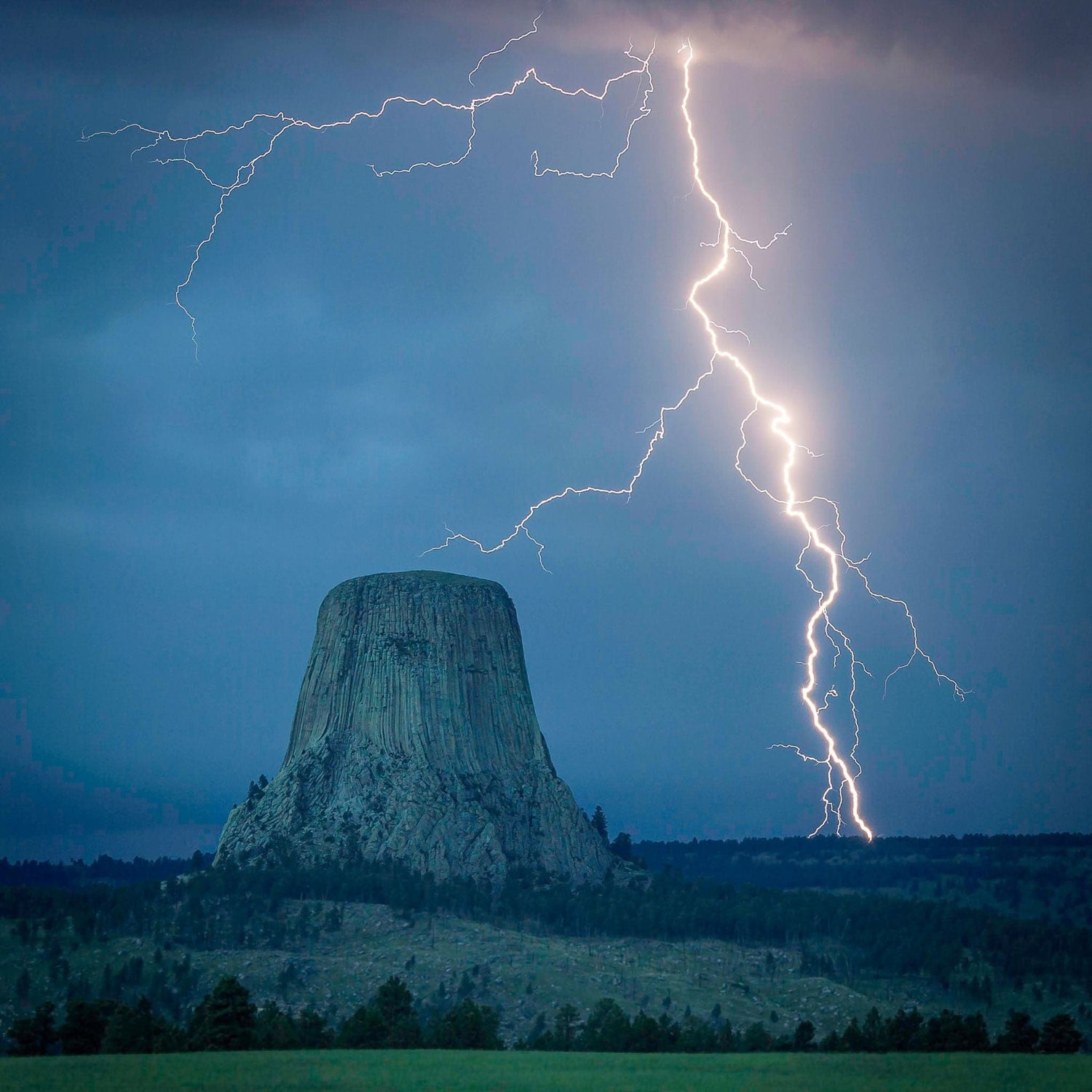 Lightning strikes near Devils Tower, a large rock formation under a dark, cloudy sky, with surrounding forested landscape.