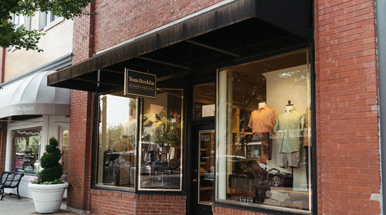 Brick storefront with black awning and sign reading Tom Beckbe, displaying clothing in large front windows.