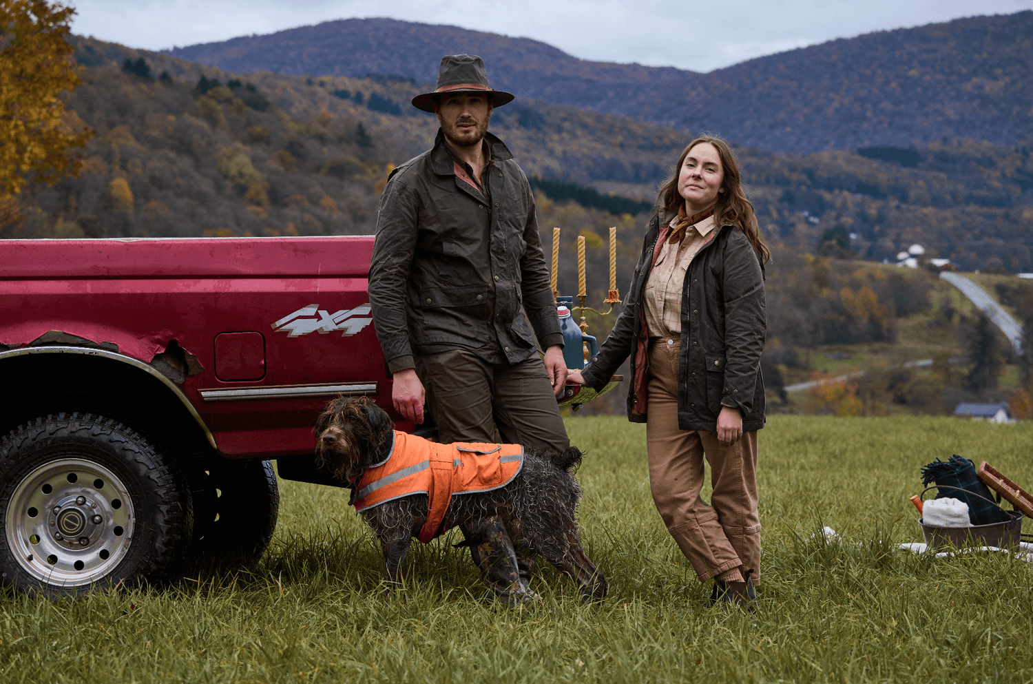 Two people in outdoor gear stand by a red truck with a dog in a field, surrounded by autumnal hills and trees.