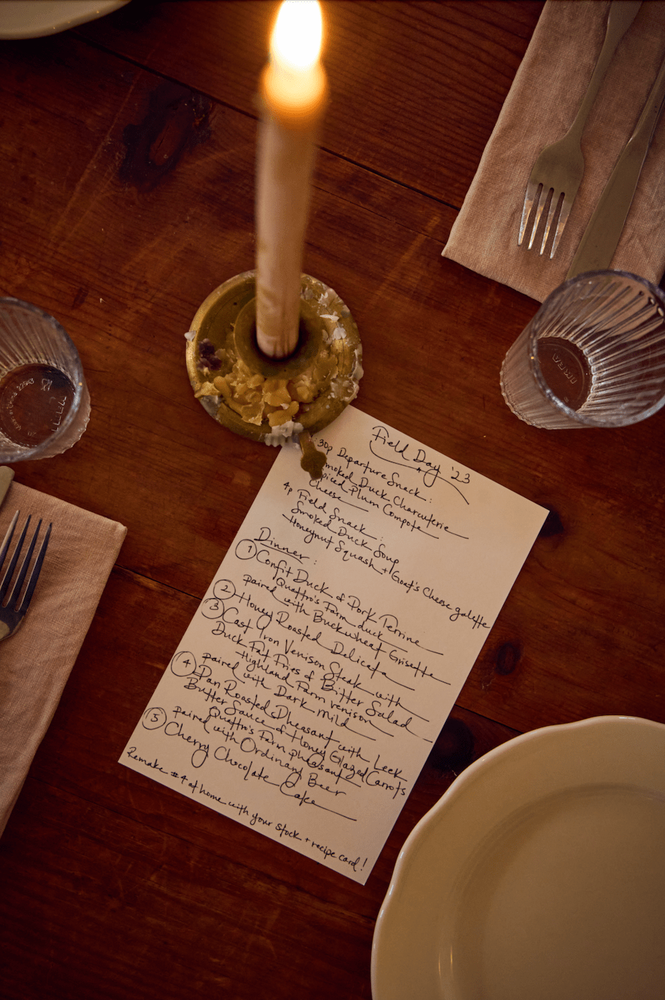 A dimly lit wooden table set with plates, utensils, and a handwritten menu. A lit candle is at the center.