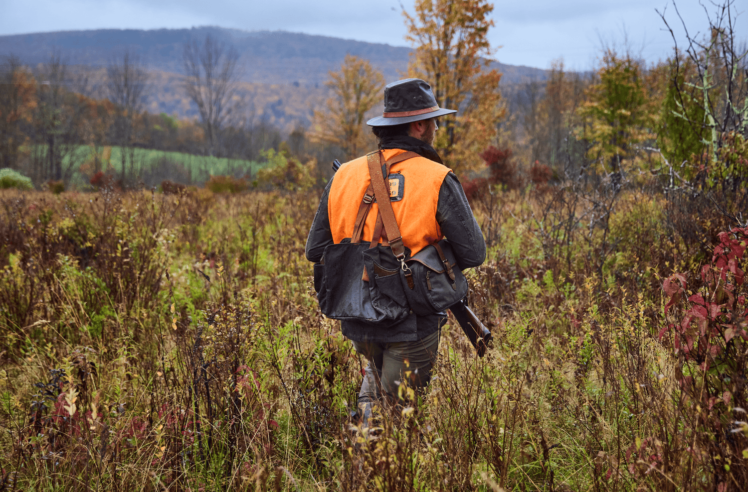 Person wearing an orange vest and hat walks through tall autumn grass in a rural field, carrying a bag and a rifle.
