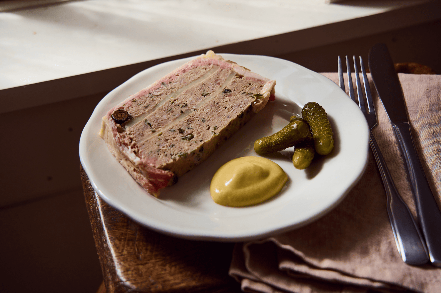 A slice of terrine with mustard and pickles on a plate, beside cutlery on a napkin, near a sunlit window.