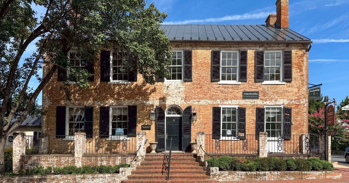 Historic two-story brick building with black shutters, arched doorway, and steps, surrounded by trees and greenery.