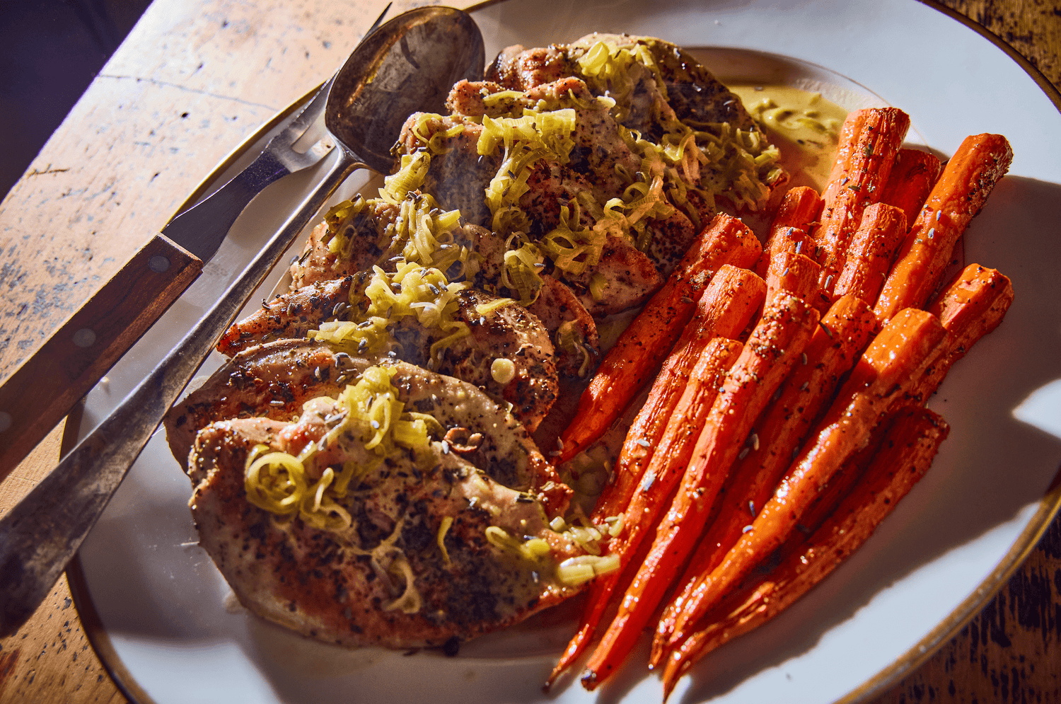 Grilled chicken topped with sliced leeks, accompanied by glazed carrots, served on a white platter with a knife and spoon.