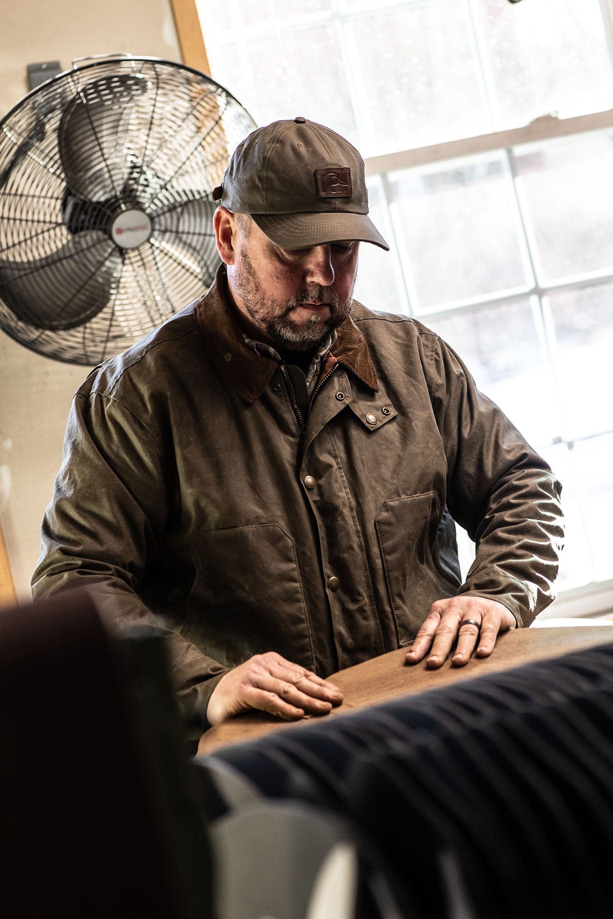 A man in a brown jacket and cap stands indoors, working with his hands on a wooden table near a window and fan.