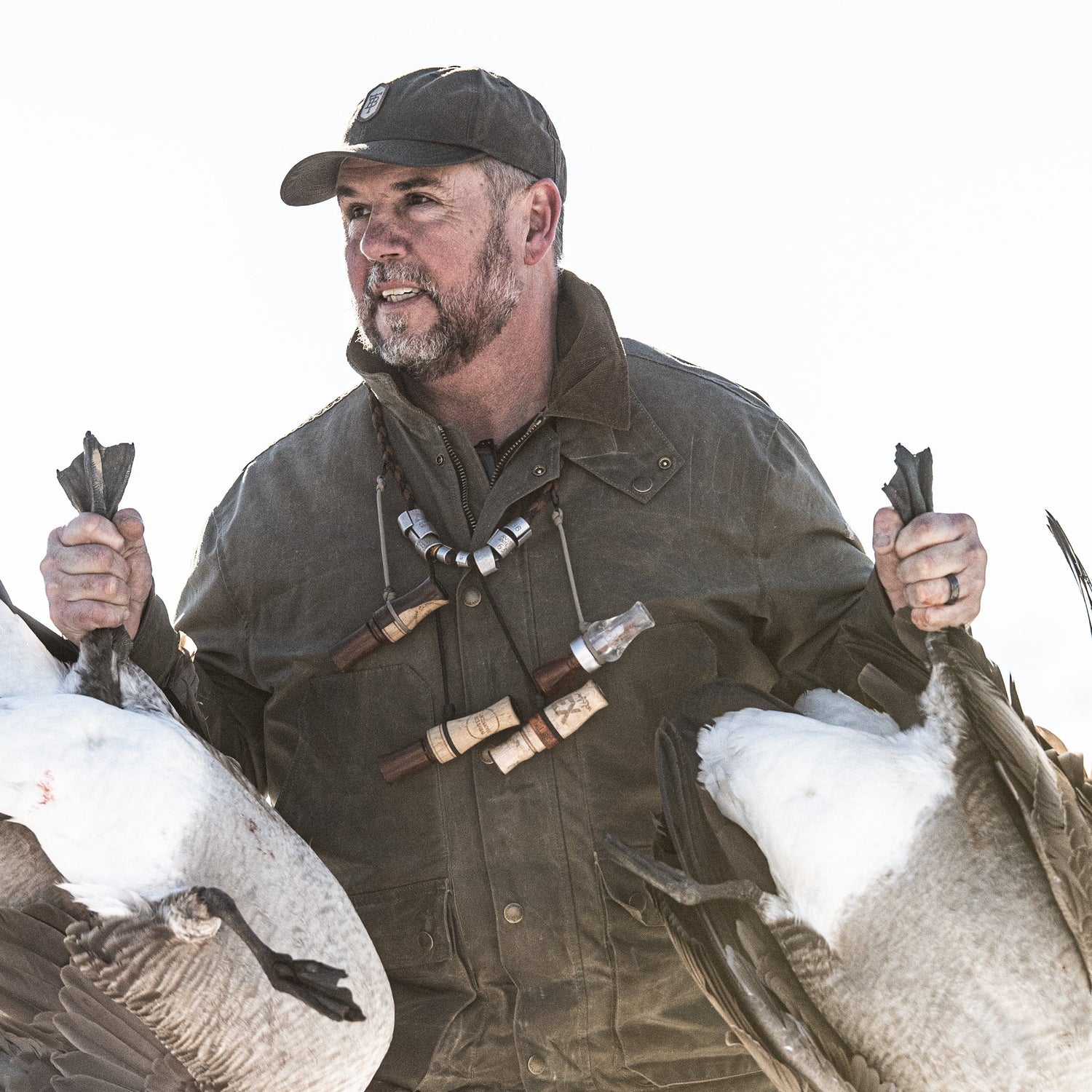 A man in outdoor gear holds two large birds by their feet, with duck calls hanging around his neck.