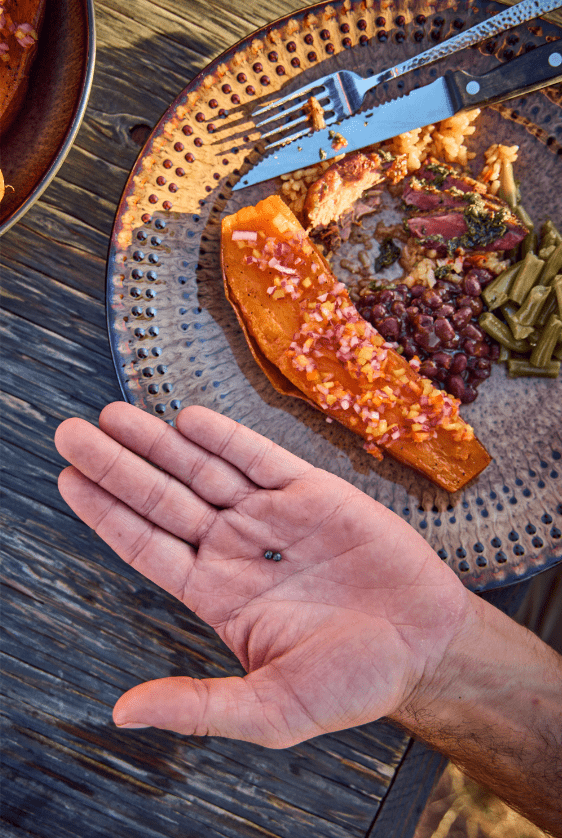 A hand holding a small black seed above a plate with a half-eaten sweet potato, beans, greens, and cutlery on a wooden table.