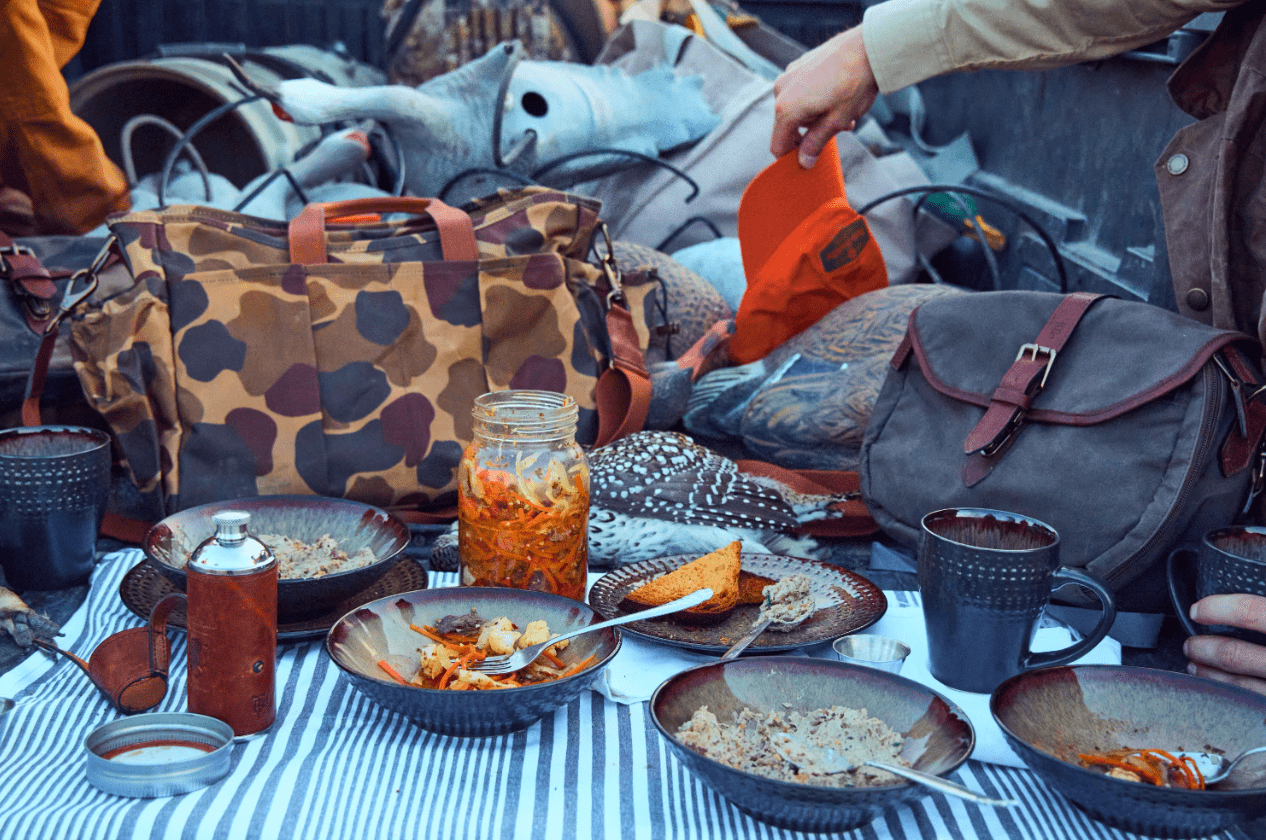Outdoor picnic scene with bowls of food, mason jar, and drinks on a striped cloth. Camouflage and grey bags are in the background.