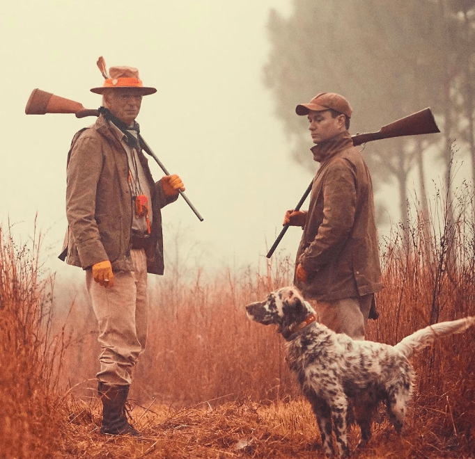 Two hunters with rifles and a dog stand in a grassy field, dressed in outdoor gear, under a misty sky.