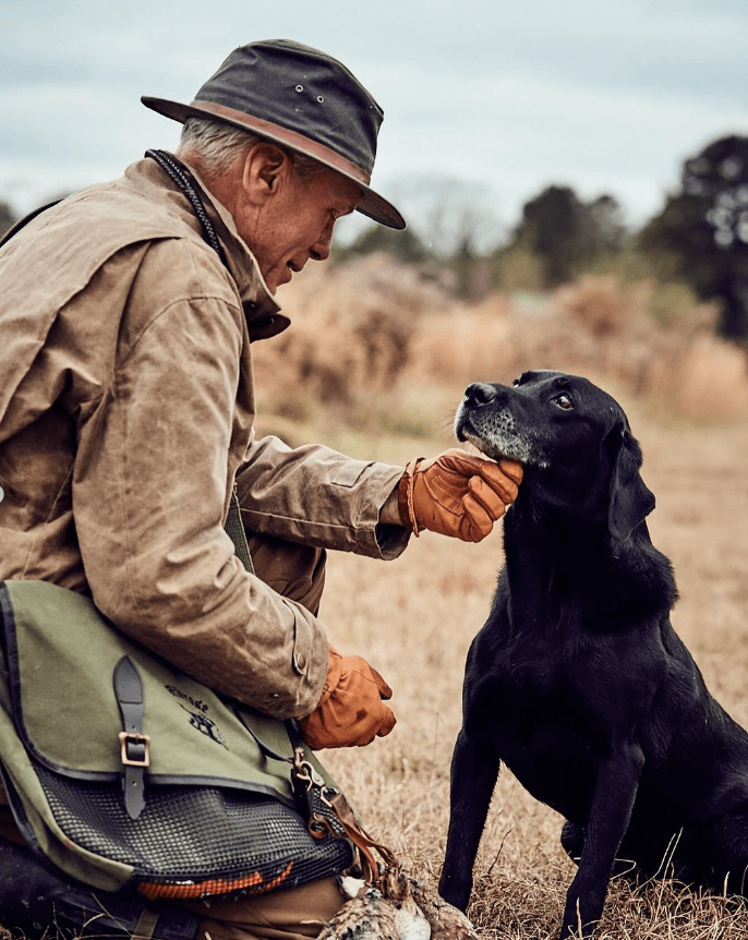 Man in a hat kneeling and petting a black dog in a grassy outdoor setting.