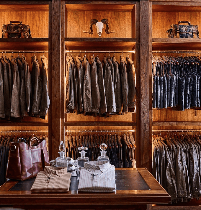 Clothing store display featuring rows of jackets and jeans on wooden shelves, a table with folded shirts and bags in the foreground.