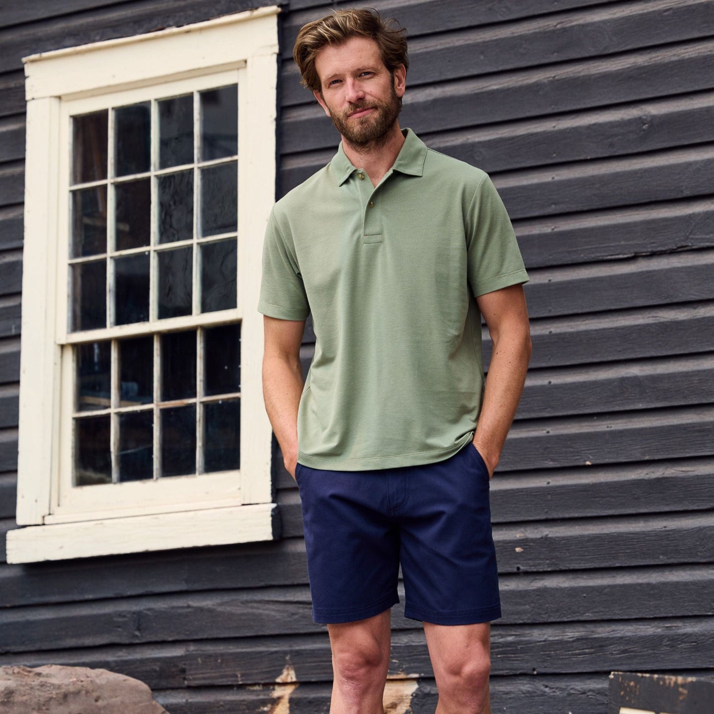 A man in a Tom Beckbe Coastal Polo Shirt and navy shorts stands by a wooden building with a white-framed window.