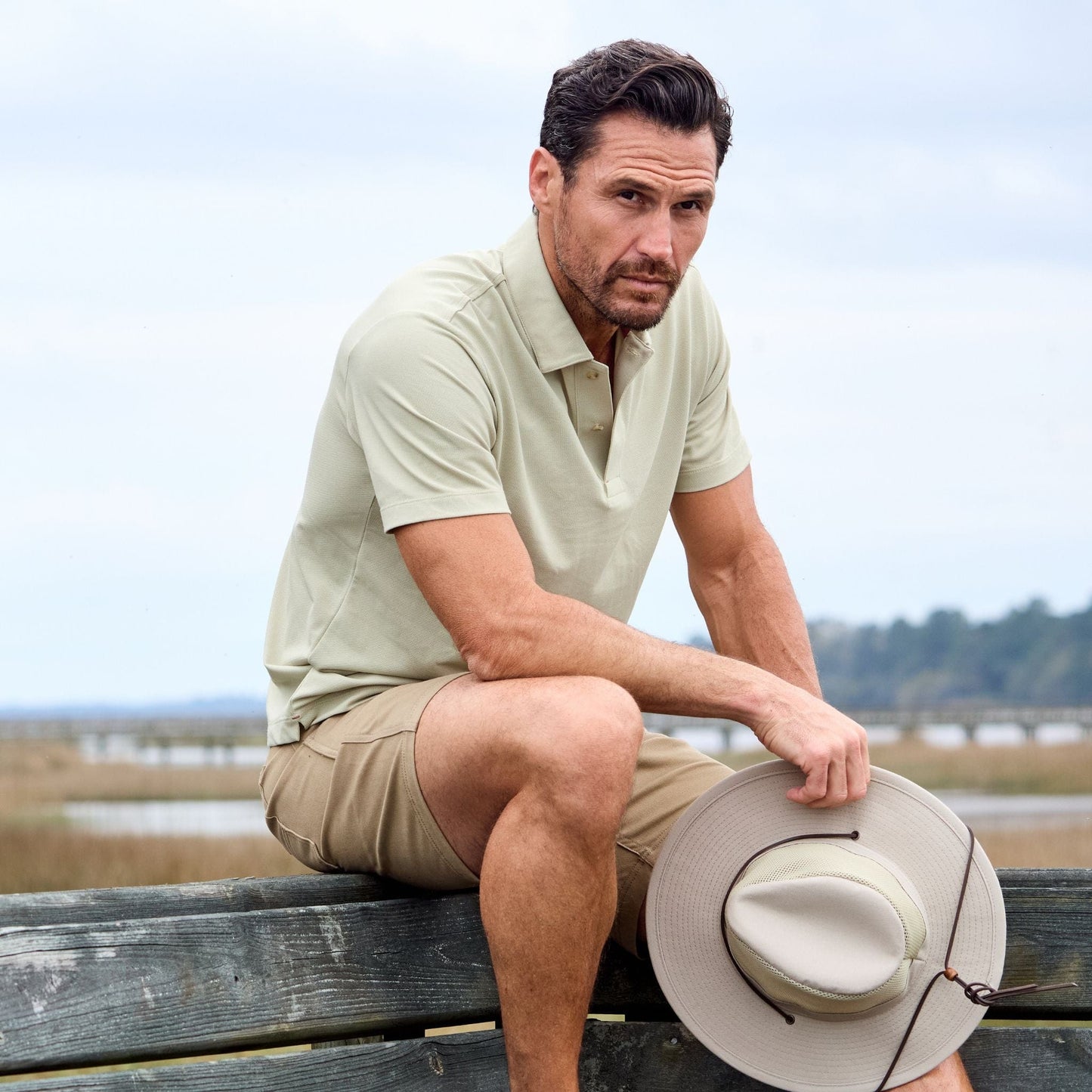 A man in a Tom Beckbe Coastal Polo and khaki shorts sits on a wooden fence with a wide-brimmed hat, marshland behind.
