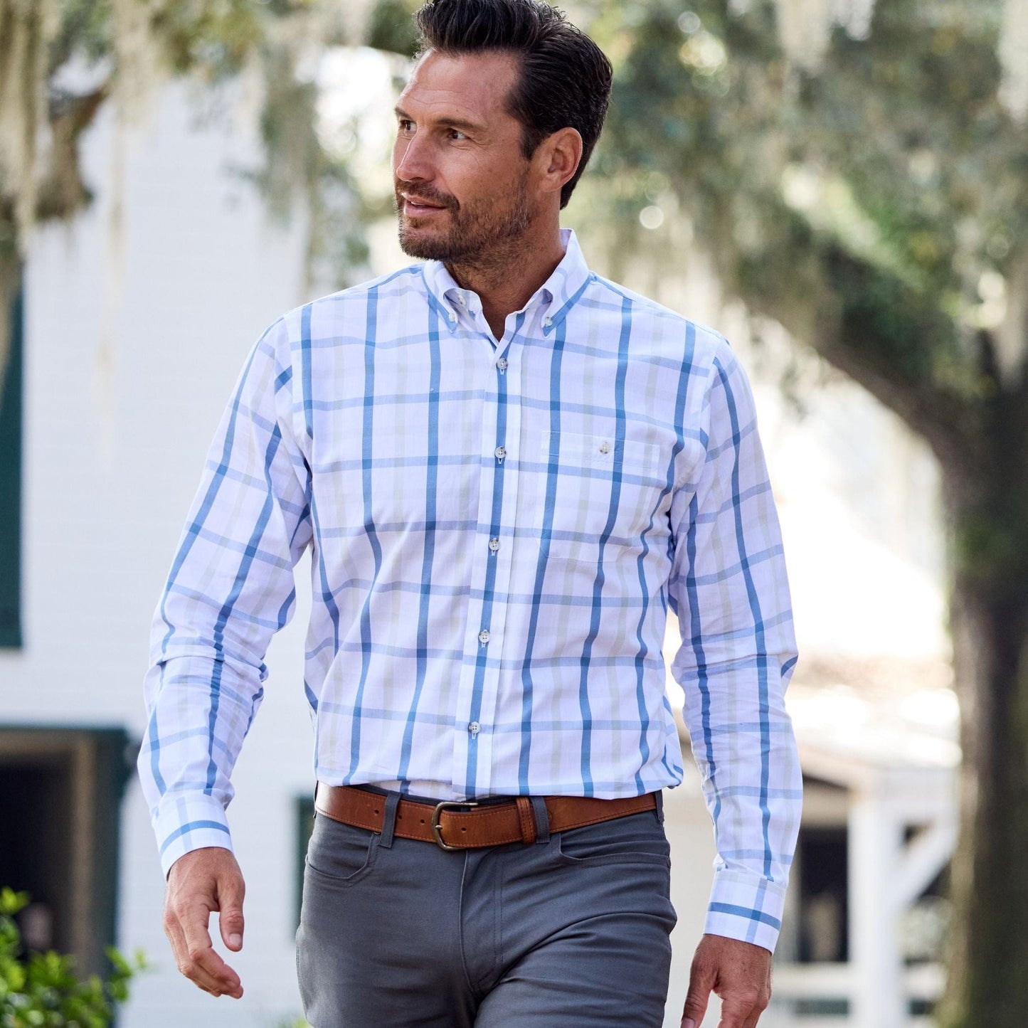 A man in a Monroe Poplin Shirt by Tom Beckbe walks outdoors, glancing sideways with trees and a building nearby.
