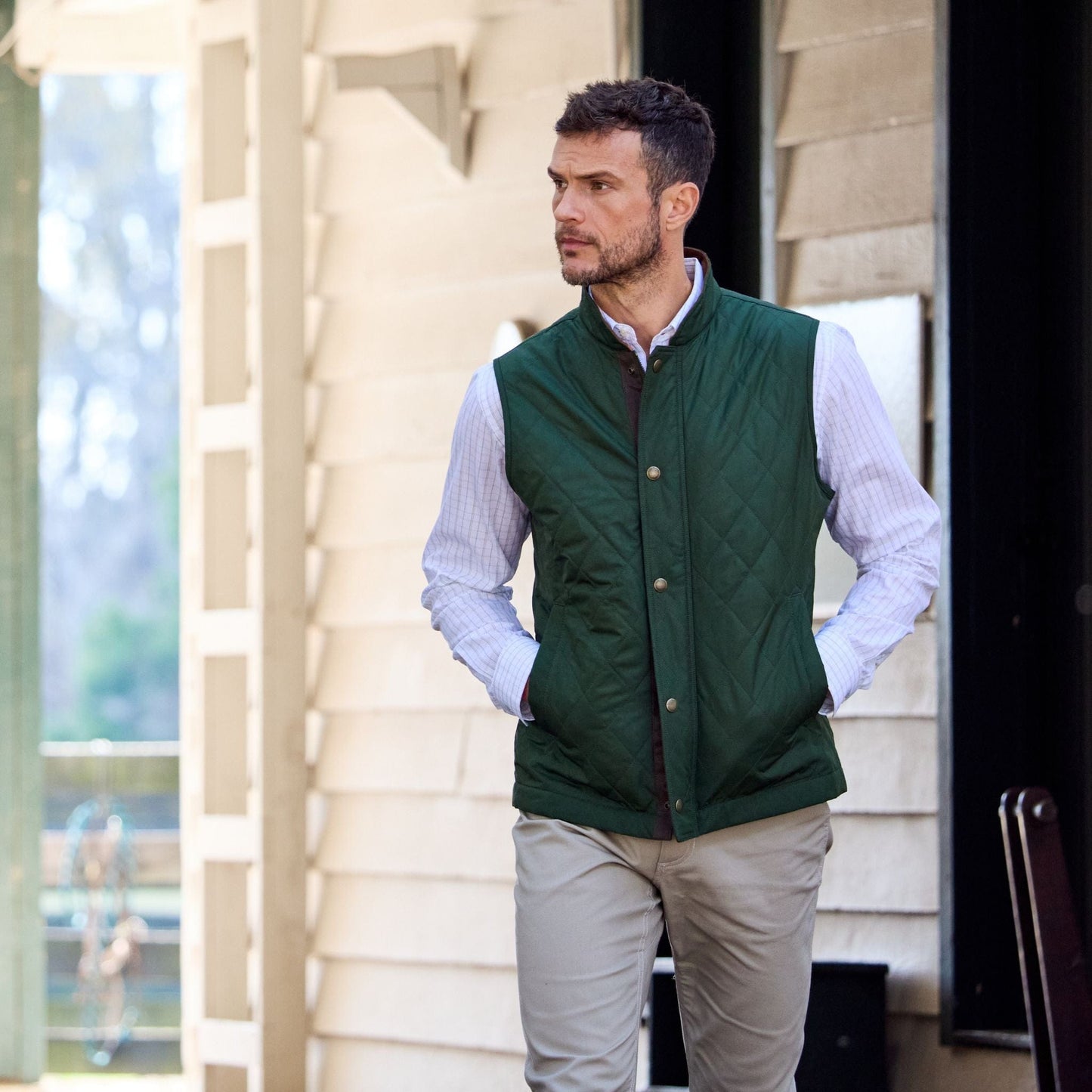 A man outdoors in a green quilted vest and Tom Beckbes Monroe Poplin Shirt, hands in pockets, glances sideways.