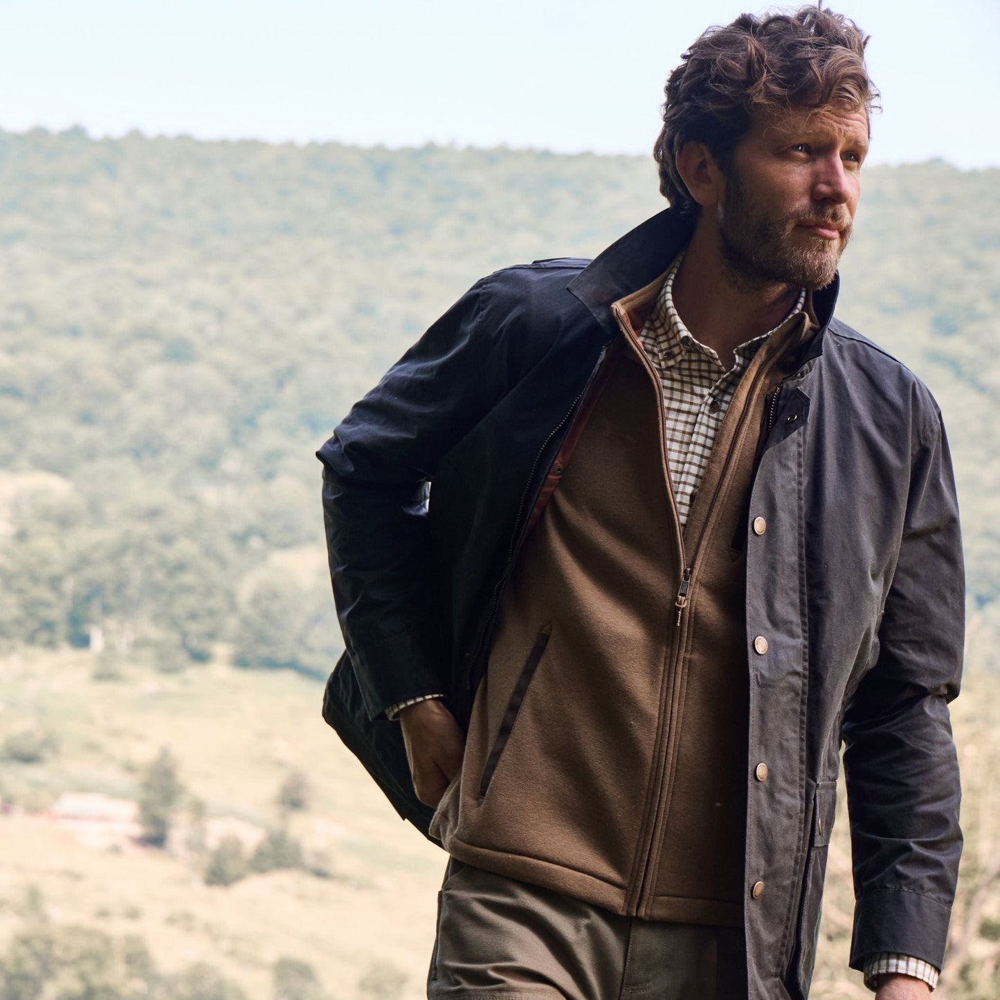 A man stands outdoors, hands in pockets, wearing a Tom Beckbe Piedmont Jacket over a sweater, with a forest backdrop.