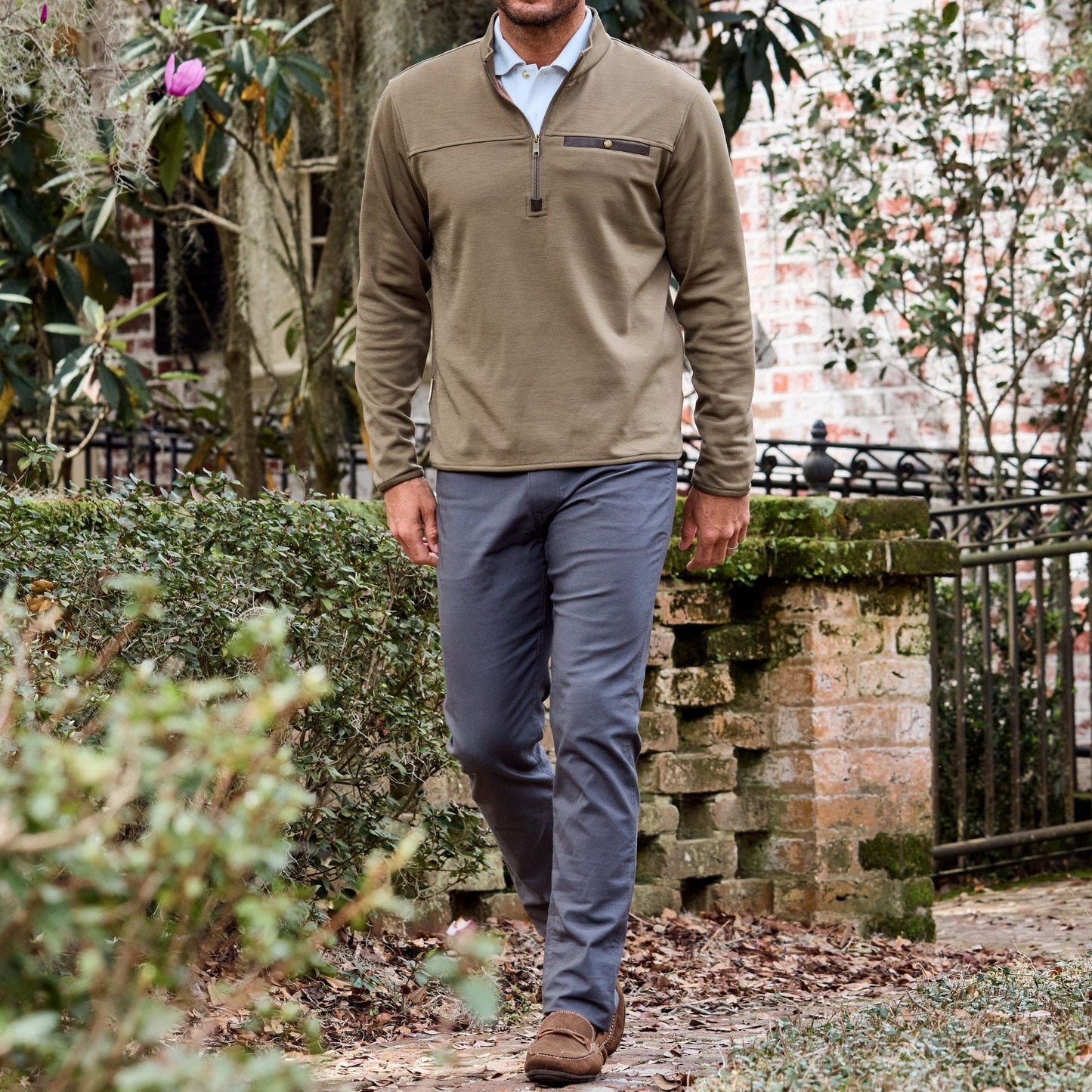 A man in a Tom Beckbe zip-up sweatshirt and Redmont Pant walks past greenery and brickwork, blurred in the background.
