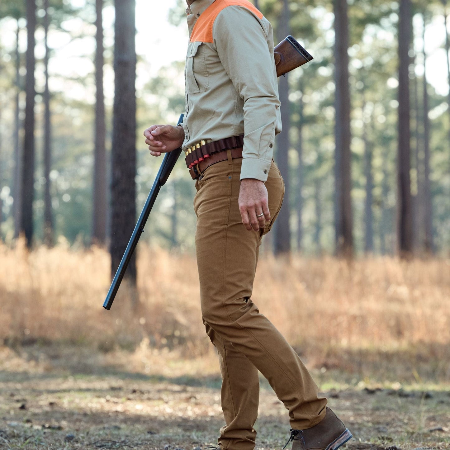 A person walks through the woods with a rifle, wearing an orange-patched beige shirt and Tom Beckbe Redmont Pant in durable cotton.