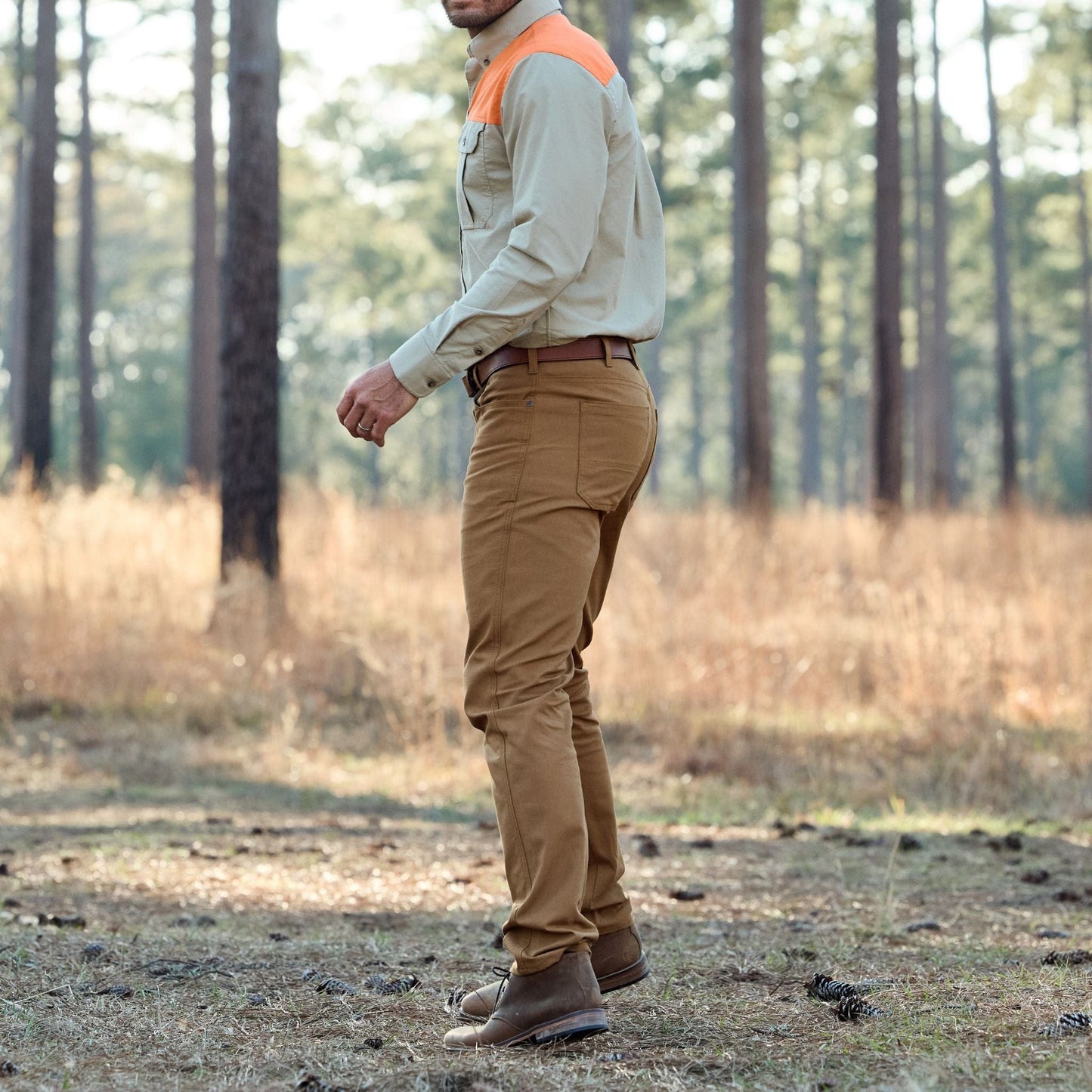 Tom Beckbes Redmont Pant and an orange-accented shirt are worn by a person standing in a forest among trees and dry grass.