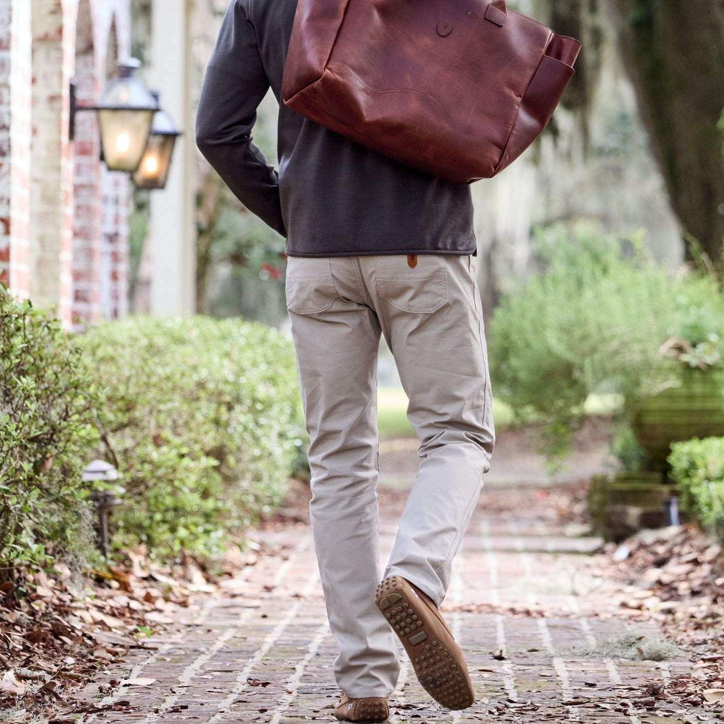 A person in Redmont Pants walks a brick path with a cotton bag, surrounded by greenery and lamps.