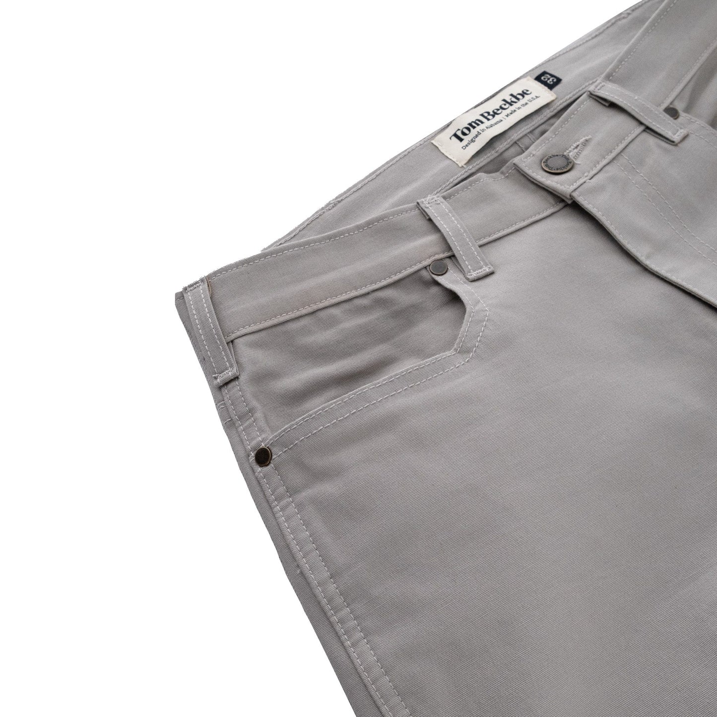 Close-up of light gray Tom Beckbe Redmont Pant, showing brand label, belt loops, durable cotton, and metal rivets.