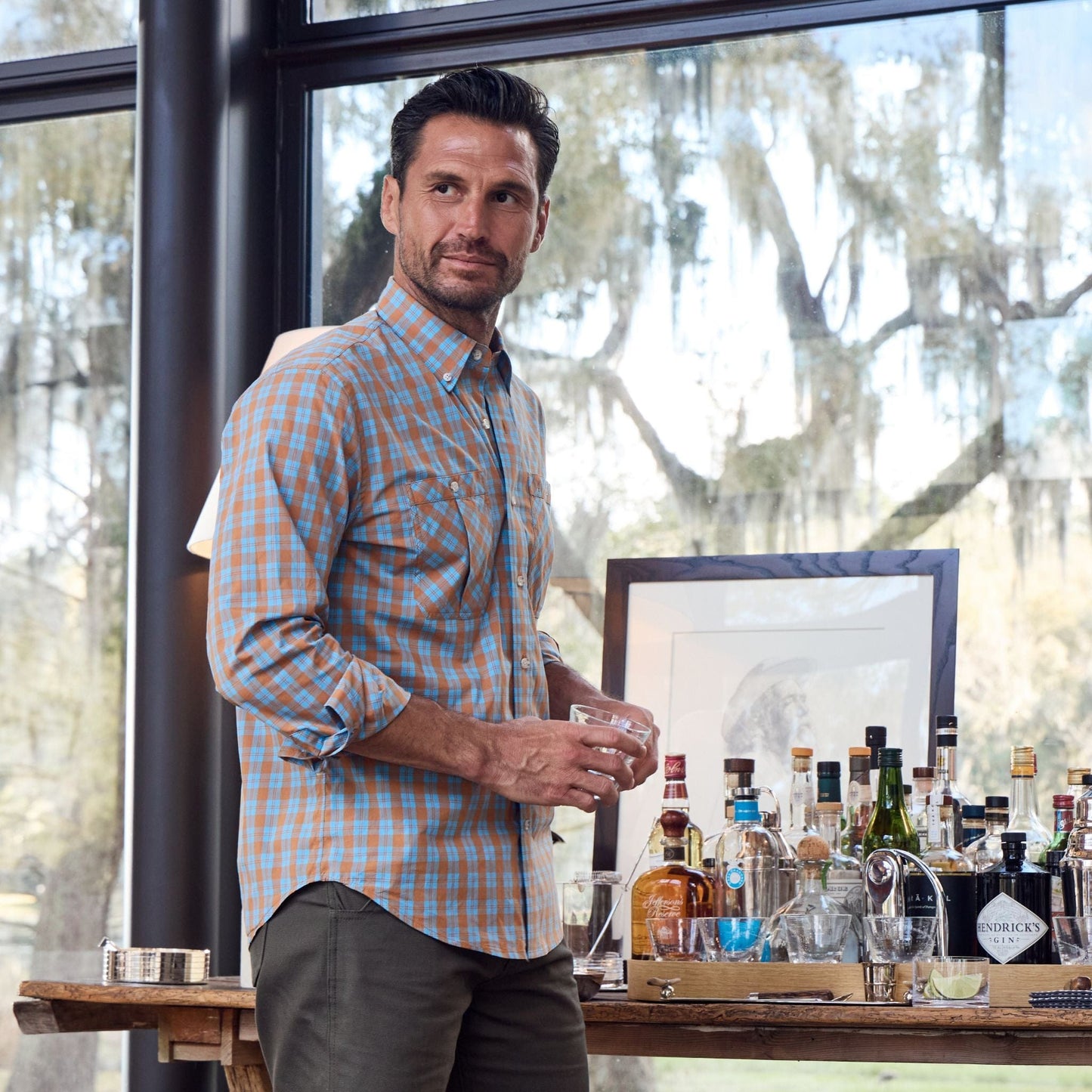 A man in a Tidewater Shirt by Tom Beckbe holds a drink beside a tray of bottles, with a large window behind him.
