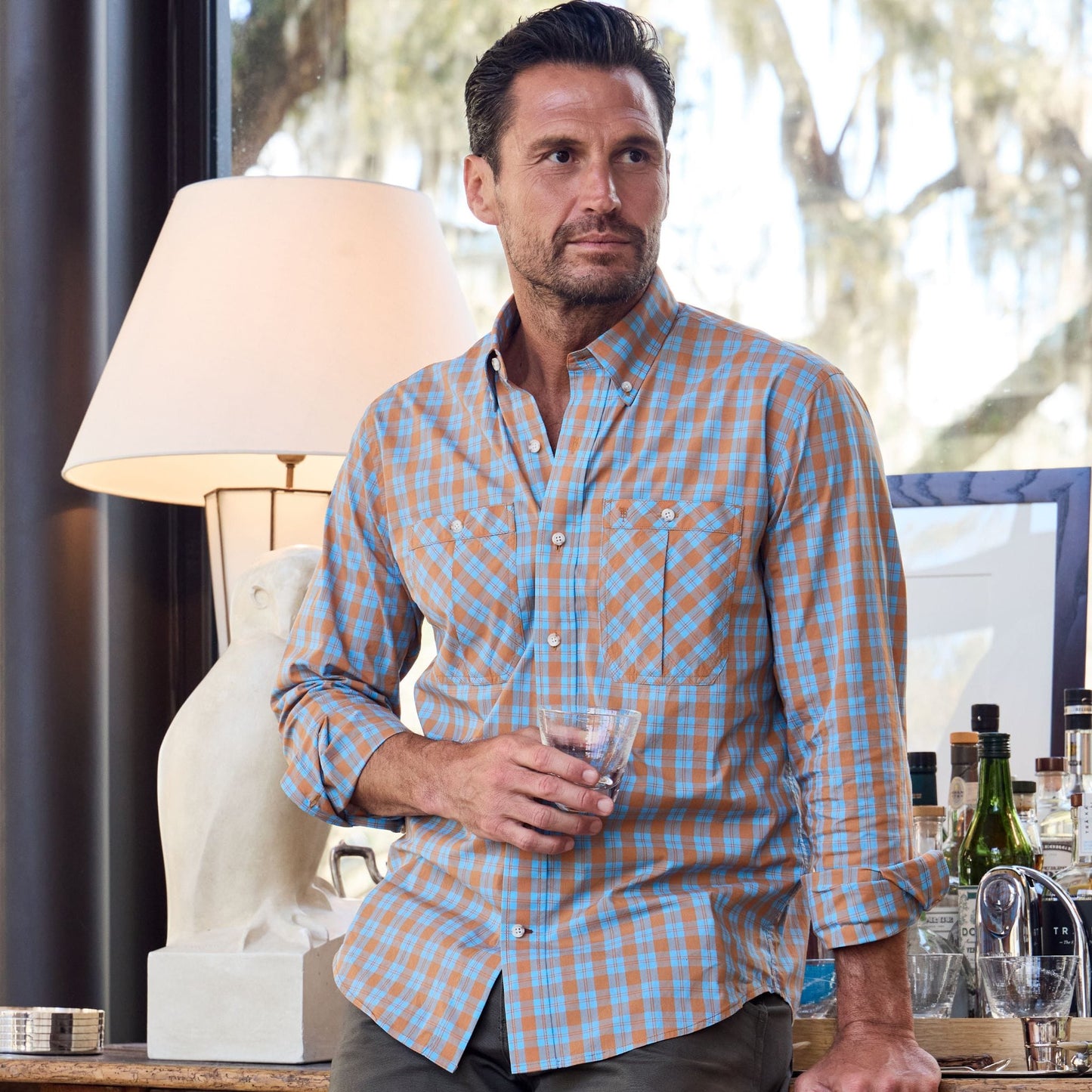 Man in a Tom Beckbe Tidewater Shirt, holding a drink, leans on a bar with bottles, lamp, and a sculpture.