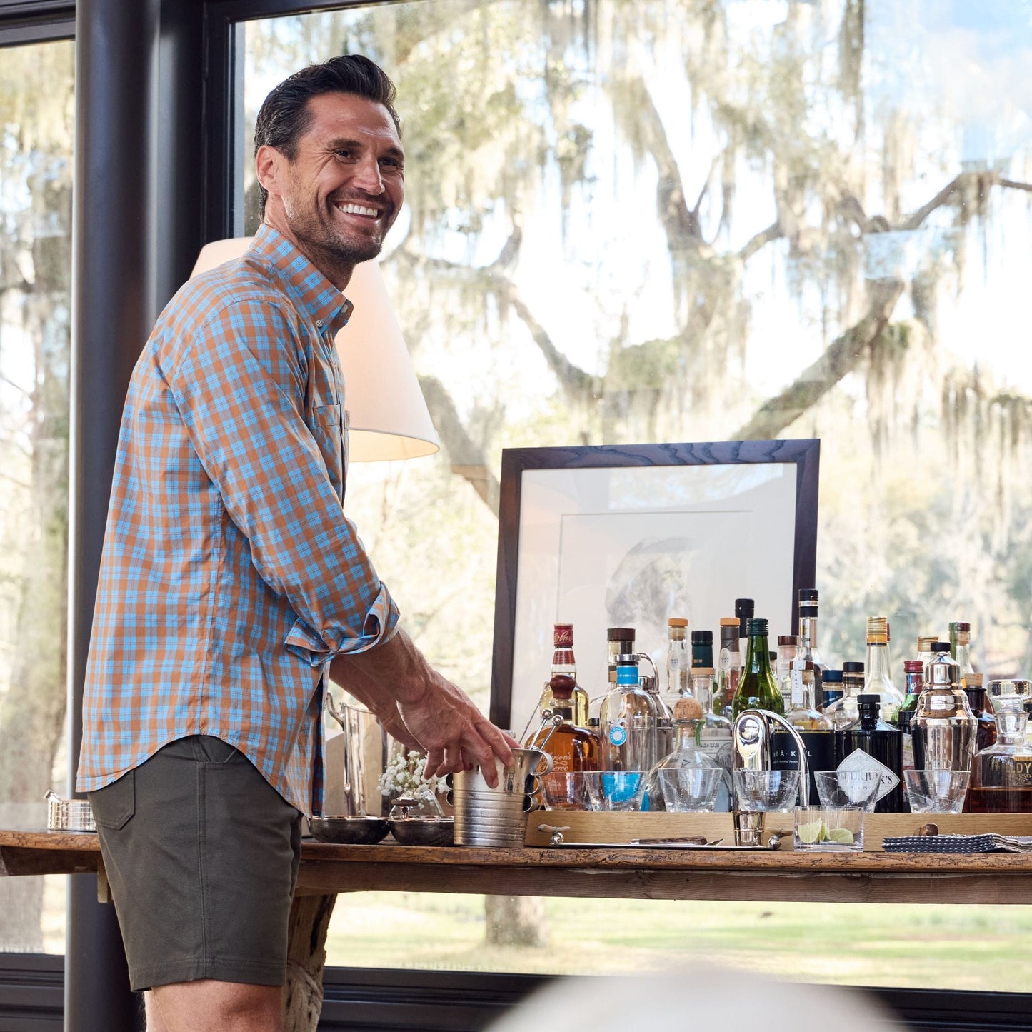 A man in a Tom Beckbe Tidewater Shirt grins by a home bar, amidst glassware and bottles near a large window.
