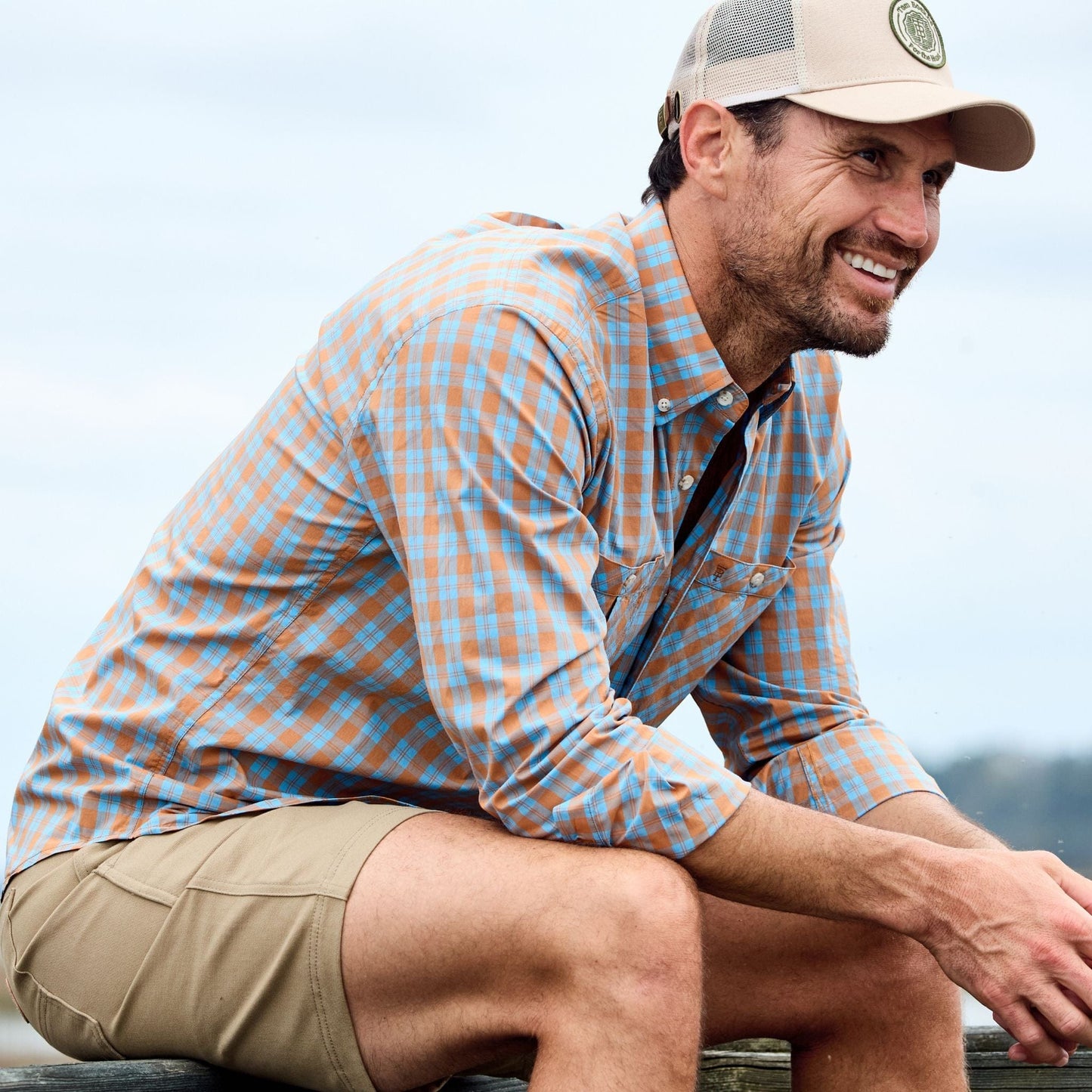 Man in a Tom Beckbe Tidewater Shirt and cap smiling outdoors wearing khaki shorts against a blurred sky background.