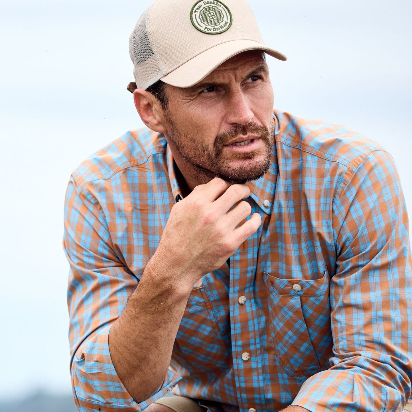 Man in Tom Beckbe Tidewater Shirt, beige cap, gazes thoughtfully to the side, resting his chin on his hand.