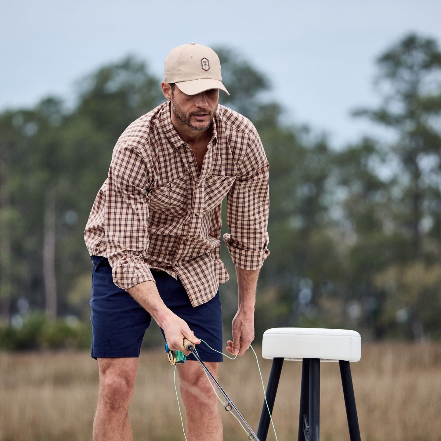 Man in a Tom Beckbe Tidewater Shirt and cap, handling a line near a stool outdoors with trees in the background.