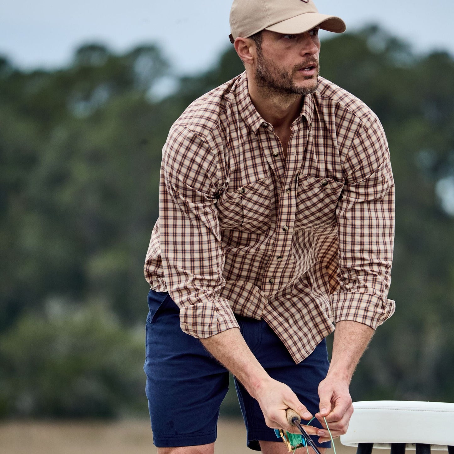 Man in a checked Tidewater Shirt by Tom Beckbe, wearing a cap and holding gear against a blurred backdrop of trees and sky.