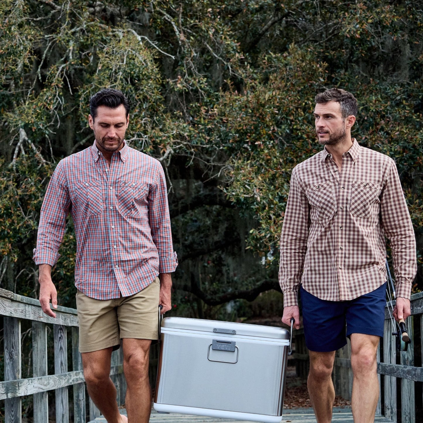 Two men in casual clothes carry a silver box on a wooden path, wearing Tom Beckbe Tidewater Shirts, surrounded by trees.