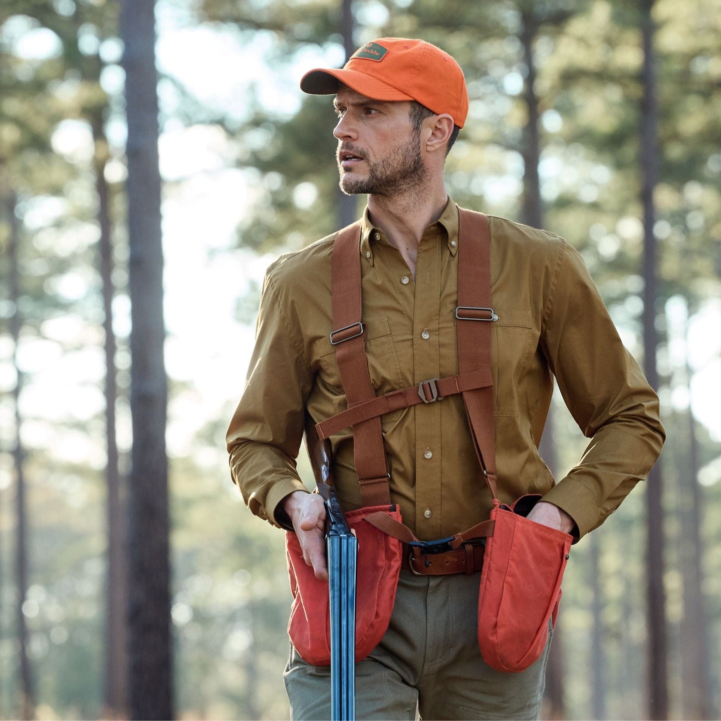 A man in an orange cap and Tidewater Shirt by Tom Beckbe walks through a forest with a rifle and UPF 30+ harness.