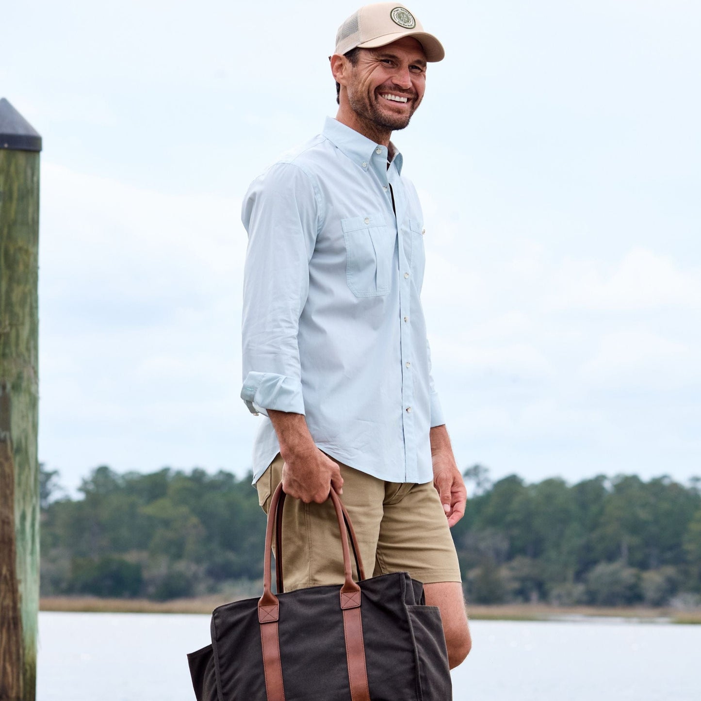 A man in a Tidewater Shirt, khaki shorts, and cap smiles by the waterfront with a dark tote bag.