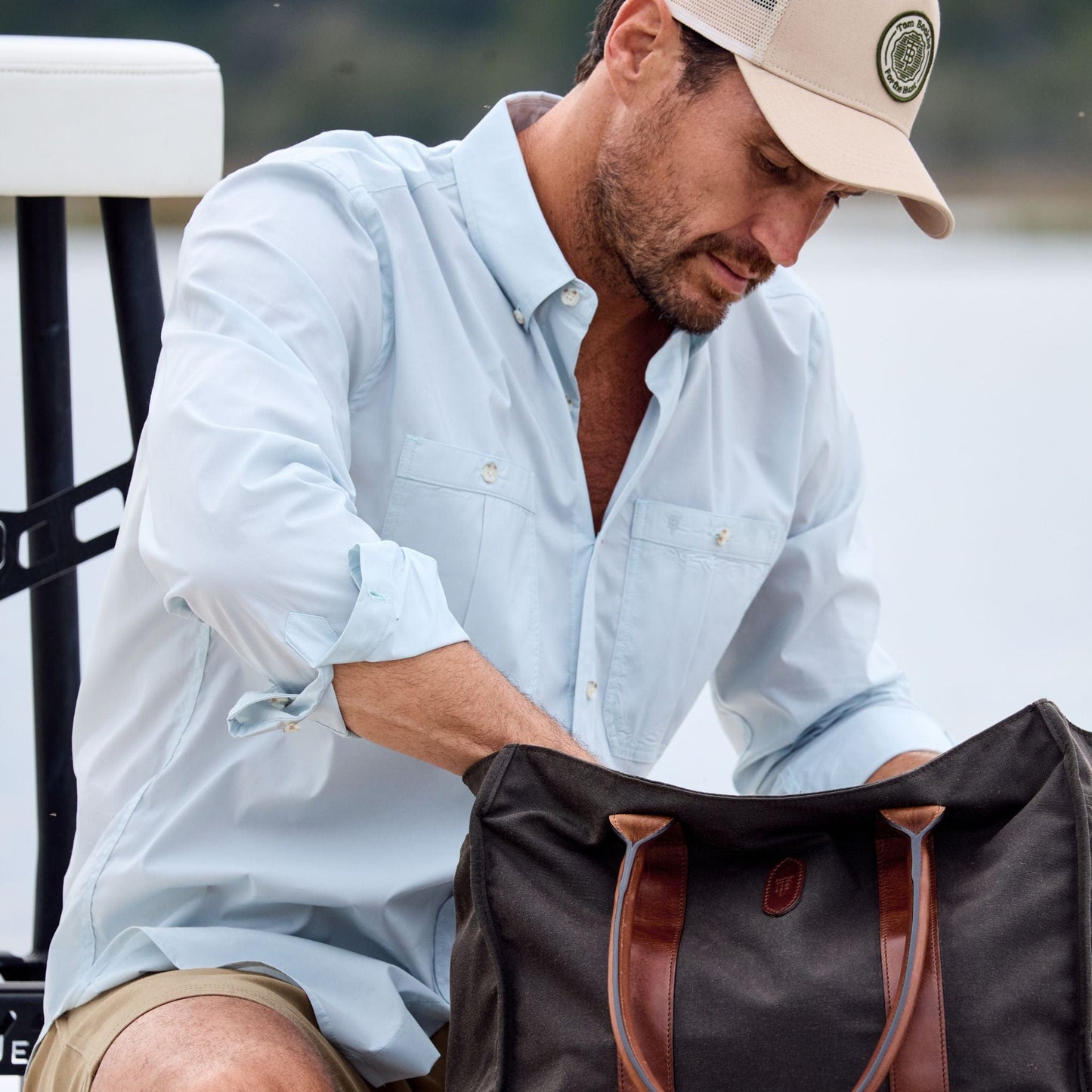 A man in a Tom Beckbe Tidewater Shirt and cap looks into a bag with brown straps.