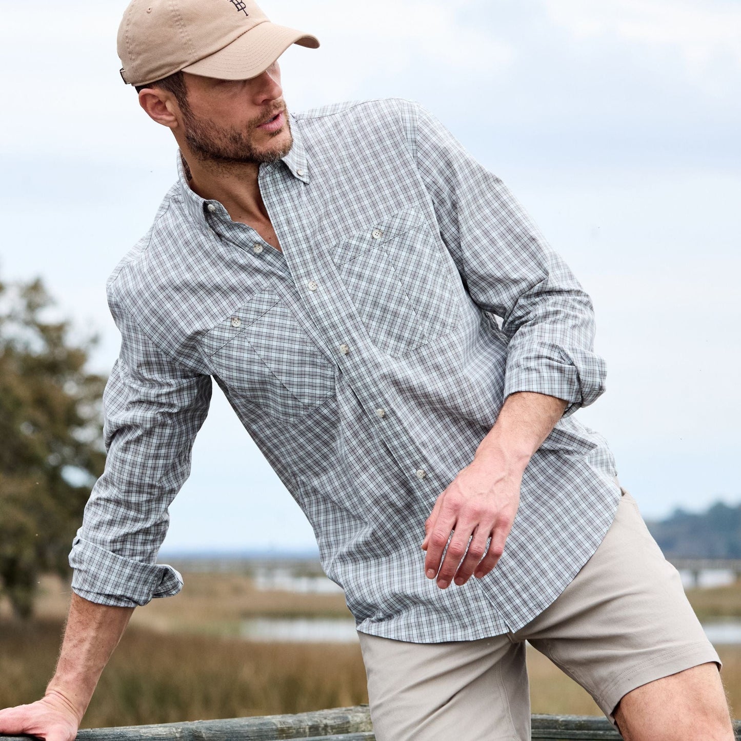 A man in a plaid Tidewater Shirt by Tom Beckbe leans against a railing outdoors, wearing a beige cap.