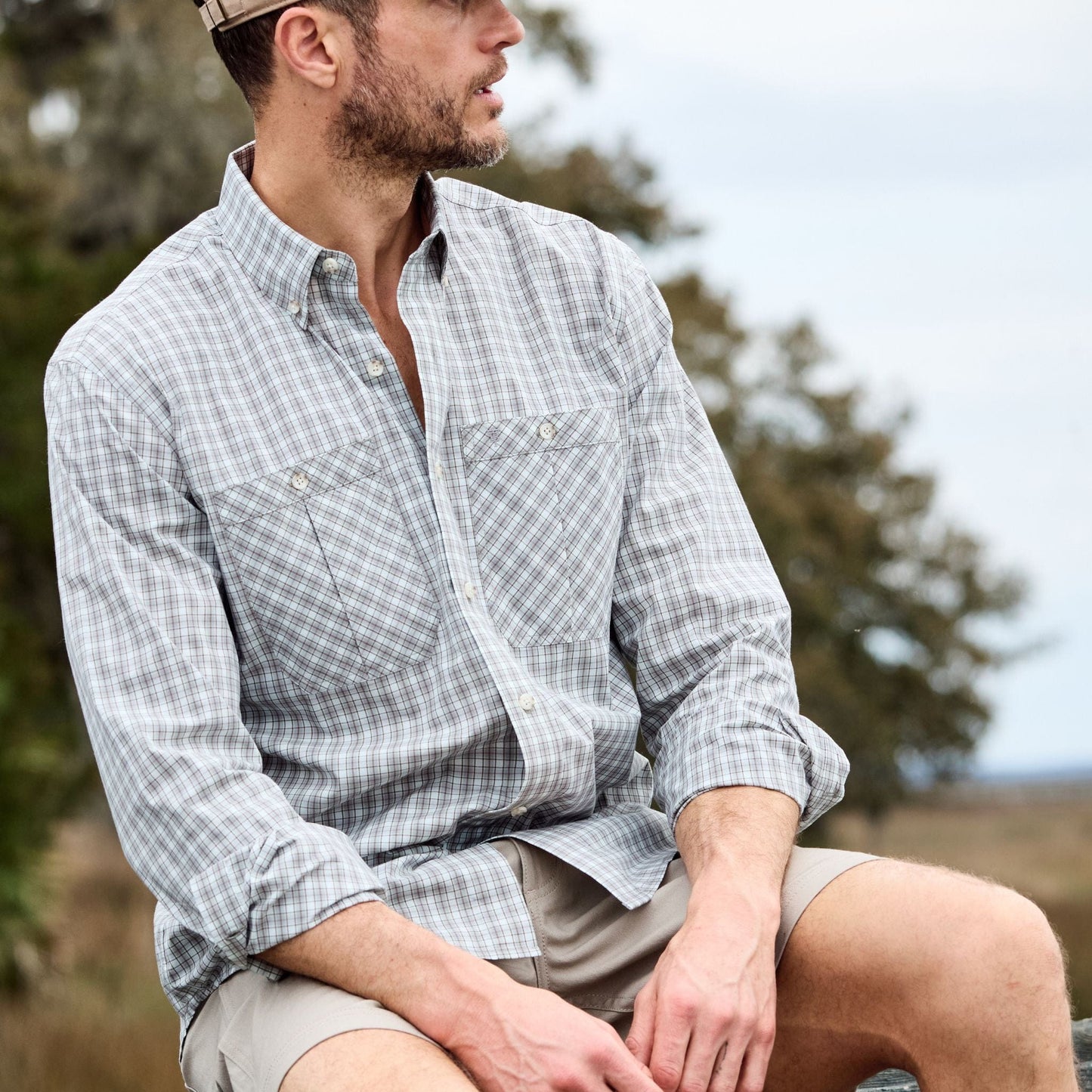A man in a Tom Beckbe Tidewater Shirt and shorts sits outside, gazing sideways, surrounded by trees and grass.